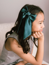 Load image into Gallery viewer, Young girl with a large teal bow in her hair, sitting on a white surface.